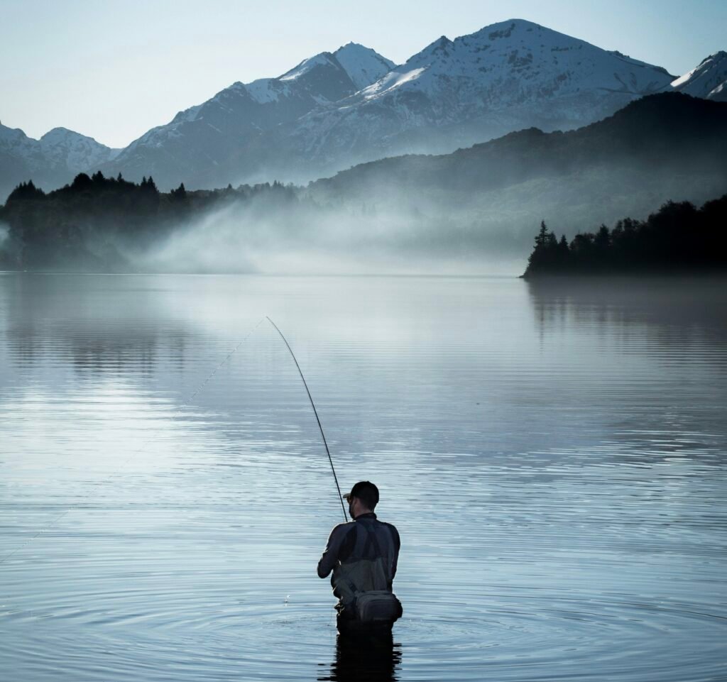 Angler fly-fishing in a misty mountain lake beneath snow-covered peaks.